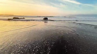 Gün batımında sakin ve sakin bir sahil, yumuşak dalgalar ve güzel altın renkler sergiliyor gökyüzünü aydınlatan