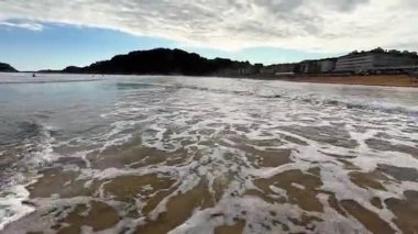 Güzel ve sakin bir sahil sahnesinde bulutlu bir gökyüzünün altında yumuşak dalgalar yavaşça kumlu kıyıya vuruyor.