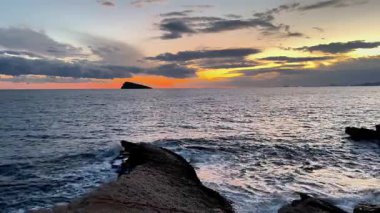 Şok edici bir günbatımı, kayalık bir kıyı şeridi ve renkli bulutlar tarafından çerçevelenmiş sakin suların üzerine canlı renkler saçar.