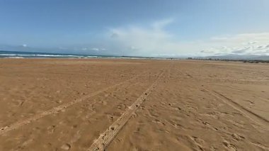 Yumuşak altın kum, yumuşak dingin dalgalar ve yukarıda canlı mavi bir gökyüzü içeren sakin bir sahil sahnesi.