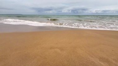 Yumuşak ve ılık kumları, kıyıya vuran hafif dalgaları ve yukarıda bulutlu bir ufku tasvir eden sakin bir sahil sahnesi.