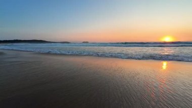 Gün batımında huzurlu ve sakin bir sahil sahnesi, hafif dalgalar ve gökyüzünde yayılan yumuşak renkler.