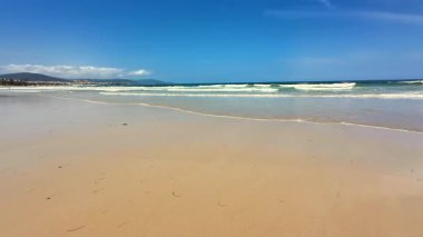 Sakin ve sakin bir plaj sahnesi yumuşak, sıcak kum, hafif dalgalar ve yukarıda açık mavi bir gökyüzü sergiliyor.