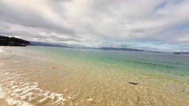 Bu sakin sahil sahnesi, bulutlu ve bulutlu bir gökyüzünün altında yumuşak kumları ve mükemmel sakin suları sergiliyor.