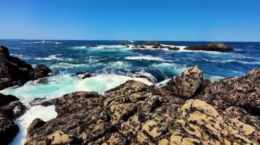 Göz kamaştırıcı bir okyanus manzarası kayalık bir kıyı şeridi, canlı dalgalar ve hepsinin üzerinde açık mavi bir gökyüzü sergiliyor.