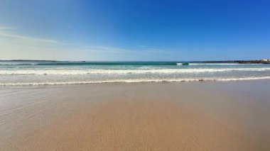 Sakin bir sahil sahnesi kumlu bir sahile ve canlı bir mavi gökyüzüne sahiptir.