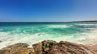 Göz kamaştırıcı bir sahil sahnesi canlı turkuaz sular ve güzel bir günde ışıldayan güneş ışığı kayaları sergiliyor.