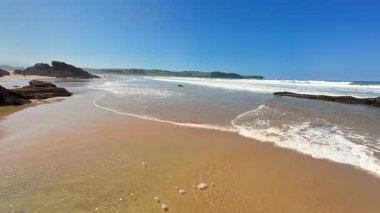 Muhteşem sahil manzaraları sakin, nazik dalgalar ve eşsiz kayalık oluşumlar sergiliyor, hepsi berrak, mavi bir gökyüzünün altında.