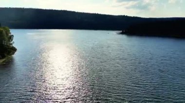 Huzurlu ve sakin bir göl, berrak mavi gökyüzünün altındaki yemyeşil tepelerin çerçevelediği güneş ışığını güzel bir şekilde yansıtır.