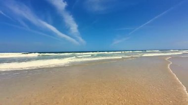 Huzurlu ve sakin bir sahil sahnesi yumuşak altın kum, berrak nazik dalgalar ve parlak mavi bir gökyüzü sergiliyor.