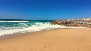 Yumuşak kum, yumuşak dalgalar ve açık bir gökyüzünün altında büyük bir kaya oluşumunun yer aldığı pitoresk bir sahil sahnesi..