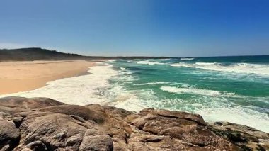 Dalgaların açık mavi gökyüzünün altındaki kayalara çarptığı güzel kumlu bir sahilin nefes kesici manzarası.