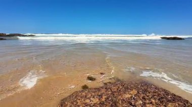 Huzurlu bir sahil sahnesi, nefes kesen berrak mavi bir gökyüzü ve yumuşak okyanus dalgalarının yavaşça aktığı.