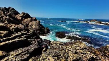 Sakin bir okyanus manzarası, parlak mavi gökyüzünün altında heybetli kayalara çarpan güçlü dalgaları gösterir.