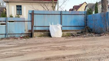 Terkedilmiş bir alanda mavi bir çit ve şehir çürümesini ve ihmalkarlığı vurgulayan büyük bir çöp yığını görülüyor.