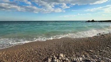 Güzel, sakin bir sahil manzarası berrak mavi gökyüzünün altında yumuşak çakıl taşlarına karşı yumuşak dalgalar sergiliyor.