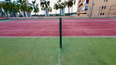 Manzaralı bir tenis kortu uzun palmiye ağaçları ile çevrilidir. Canlı kırmızı yapay çim oyun için idealdir.