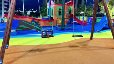 Salıncaklar, slaytlar ve gece renkli manzaralarla çevrili canlı ve hareketli bir oyun alanı.