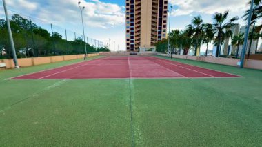 Yüksek palmiye ağaçları ve arkasında büyüleyici bir bina olan bakımlı bir tenis kortunun manzarası.