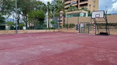 Geniş açılı çekimde geniş ağaçlarla çevrili canlı kırmızı yüzeyi olan boş bir basketbol sahası görülüyor.