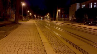Gecenin köründe sakin, ıssız bir cadde. Yumuşak parlayan sokak lambaları ve farklı tramvay rayları sergileniyor.