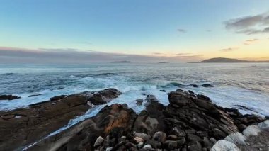 Gün batımında kayalara çarpan yumuşak dalgaların olduğu sakin bir sahil manzarası güzellikleri gözler önüne seriyor.