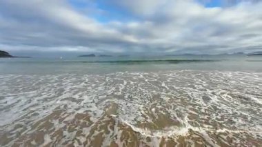 Yumuşak dalgaları ve kasvetli gökyüzünü güzel bir şekilde yansıtan sakin bir sahil manzarası, seyahat ya da doğa teması için ideal.