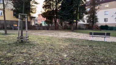 Güzel, sakin bir park sahnesi salıncaklar ve banklar sergileniyor, yemyeşil ağaçların ve yakınlardaki binaların arasında.