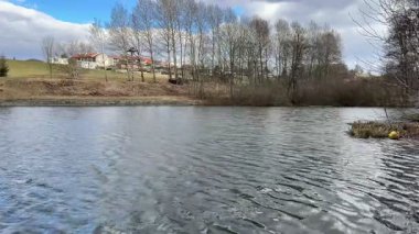 Bereketli ağaçlarla çevrili, bulutlu bir gökyüzünün altında sakin bir göl manzarası ve büyüleyici evler.