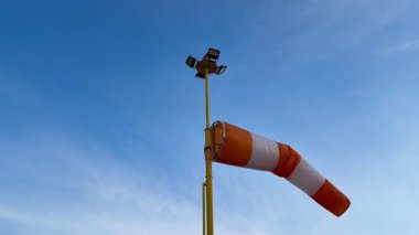 Şaşırtıcı derecede açık mavi bir gökyüzü, uzun bir direğe monte edilmiş çeşitli ışıklandırma ekipmanlarıyla birlikte renkli bir rüzgar çorabı sergiliyor.