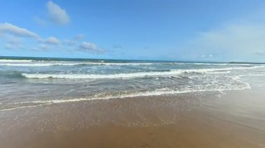 Resimli ve sakin bir sahil sahnesi, parlak mavi gökyüzünün altında süzülen nazik dalgaları sergiliyor.