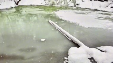 Huzurlu ve pitoresk bir kış manzarası kısmen donmuş bir su kütlesini karla kaplı bir kıyıda sergiliyor.