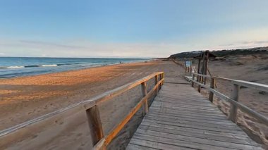 Sakin, ahşap bir patika gün batımında kumlu bir sahile çıkar. Sakin okyanus dalgaları yavaşça kıyıya vurur.