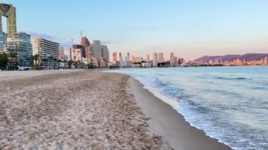 Yumuşak, yumuşak dalgaların olduğu büyüleyici sahil manzarası, pitoresk bir şehir silueti, ve güzel, sakin günbatımı renkleri