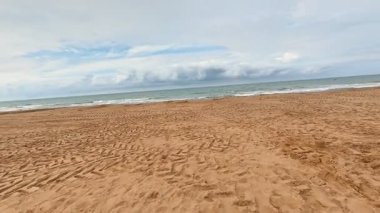 Sakin ve çarpıcı bir plaj sahnesi yumuşak altın kum, hafif dalgalar ve açık bir gökyüzü sergiliyor.