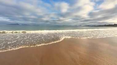 Güzel, sakin bir sahil sahnesi tasvir edilir. Nazik dalgalar ve bulutlu gökyüzü sergilenir.