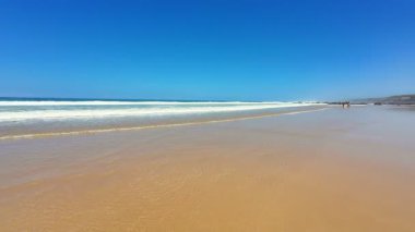 Göz kamaştırıcı bir plaj sahnesi yumuşak altın kum, kıyıya vuran nazik dalgalar ve yukarıda parlak mavi bir gökyüzü.