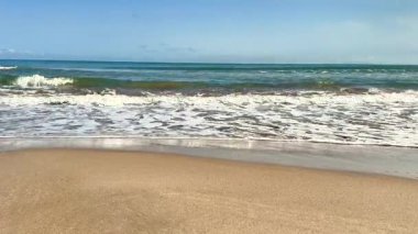 Huzurlu ve sakin bir sahil sahnesi berrak mavi gökyüzünün altında yumuşak, sıcak kum ve nazik okyanus dalgalarını sergiliyor.