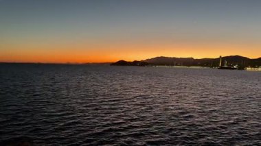 Şok edici bir günbatımı, sakin suların üzerinde gökyüzünü ve canlı bir şehrin ufkunu boyar, büyüleyici bir manzara yaratır.