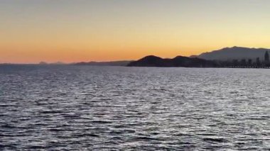 Sakin bir sahil şeridi, canlı bir şehir silueti ve çarpıcı günbatımı renkleriyle nefes kesici bir akşam manzarası.