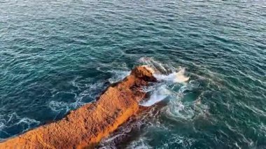 Sahilde günbatımının nefes kesici bir görüntüsü, yakınlardaki kayalık oluşumlara doğru hafif dalgalar çakıyor.