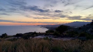Kıyı şeridinde nefes kesici bir günbatımı beliriyor. Yuvarlanan tepeler ve renkli gökyüzü tarafından çerçeveleniyor.