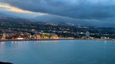 Bu çarpıcı manzara, alacakaranlıkta parlayan, doğa ve mimariyi güzelce birleştiren canlı bir şehir kıyı şeridini yansıtıyor.