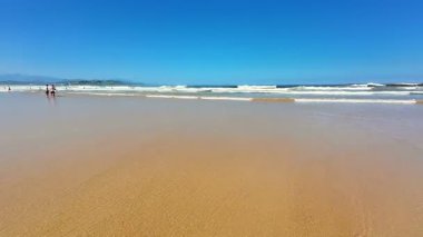 Yumuşak, altın kum, yumuşak dalgalar ve berrak mavi gökyüzü içeren sakin ve sakin bir sahil sahnesi.