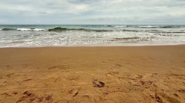 Sakin bir sahil sahnesi yumuşak dalgalarla dolu bulutlu bir gökyüzü altında altın kumları yalayıp yutan