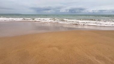 Yumuşak ve ılık kumları, kıyıya vuran hafif dalgaları ve yukarıda bulutlu bir ufku tasvir eden sakin bir sahil sahnesi.