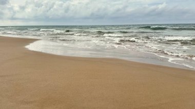 Sakin bir sahil sahnesi yumuşak dalgaları sergiliyor. Açık bir gökyüzünün altında sıcak, kumlu sahile hafifçe çarpıyor.
