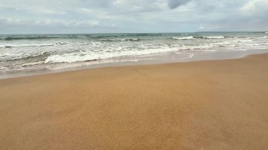 Bulutlu bir gökyüzünün altında yumuşak, altın kum ve nazik okyanus dalgalarının olduğu huzurlu ve huzurlu bir sahil sahnesi.