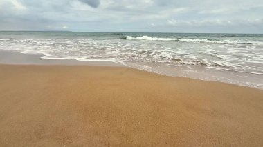 Sakin ve sakin bir sahil sahnesinde bulutlu bir gökyüzünün altında yumuşak dalgalar yavaşça kumlu kıyıya vuruyor.