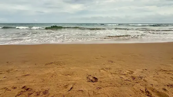 Sakin bir sahil sahnesi yumuşak dalgalarla dolu bulutlu bir gökyüzü altında altın kumları yalayıp yutan
